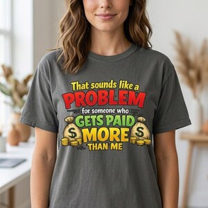 May include: A charcoal gray t-shirt featuring the text "That sounds like a PROBLEM for someone who GETS PAID MORE THAN ME" in yellow, red, and green, accompanied by illustrations of two beige money bags and stacks of gold coins.
