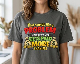 May include: A charcoal gray t-shirt featuring the text "That sounds like a PROBLEM for someone who GETS PAID MORE THAN ME" in yellow, red, and green, accompanied by illustrations of two beige money bags and stacks of gold coins.