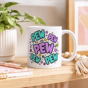 Back side of the "Pew Pew Madafakas" capybara coffee mug by SarcasmArt with colorful PEW PEW PEW lettering, on a desk with books.