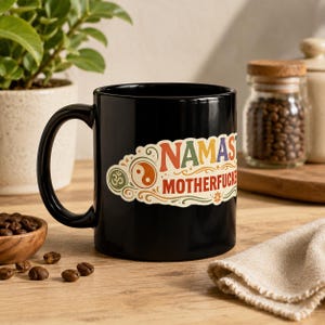 "Namaste Motherfuckers" funny yoga coffee mug by SarcasmArt: glossy black ceramic mug with a colorful boho mandala label with the adult-humor phrase NAMASTE MOTHERFUCKERS in red, orange and teal, on a wooden table with coffee beans and a plant.