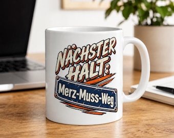 Op de afbeelding: Een witte keramische mok met een grafisch ontwerp met de tekst NÄCHSTER HALT: Merz-Muss-Weg. Het ontwerp bevat oranje penseelstreken en een blauw rechthoekig bord met witte letters, dat lijkt op een bord van een halte.