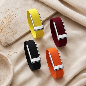 May include: Four elastic wristbands in yellow, burgundy, black, and orange. Each band features a rectangular silver-toned metal clasp with a small engraved letter W. The bands have a smooth, matte texture and are arranged in a circular formation.