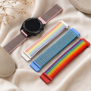 May include: A black smartwatch with a pink woven nylon strap, accompanied by three additional replacement watch bands: one with a white and rainbow stripe pattern, one in solid blue, and one with a vibrant full-spectrum rainbow gradient.