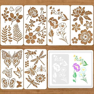 May include: A set of eight rectangular white stencils featuring cut-out designs of various botanical elements including leaves, ferns, roses, poppies, butterflies, bees, dragonflies, and a guide showing colored floral patterns on a white background.
