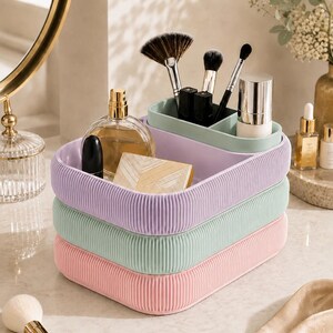 May include: A stack of three rectangular cosmetic organisers with rounded corners and a vertical ribbed texture. The top tray is lavender with a sage green insert, the middle tray is sage green, and the bottom tray is light pink. The trays hold makeup brushes, perfume, and cosmetics.