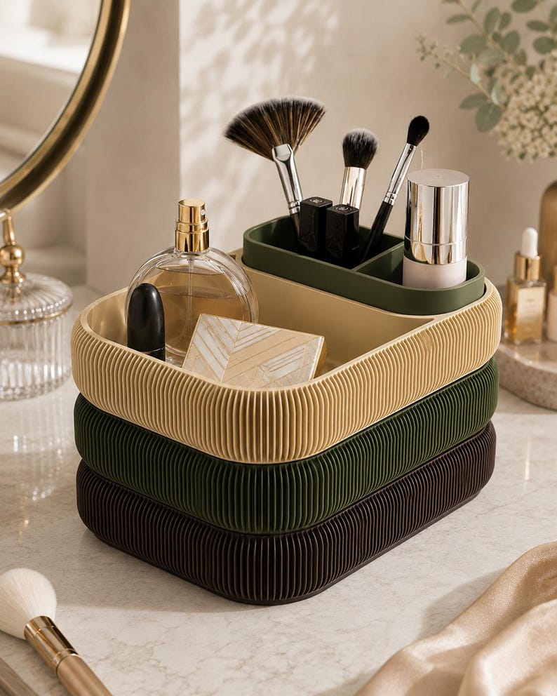 May include: A stack of three oval-shaped storage trays with a ribbed exterior texture. The top tray is beige, the middle is dark green, and the bottom is dark brown. The top tray holds a glass perfume bottle, a lipstick, and makeup brushes.