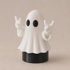 Puede incluir: Una figura de fantasma blanca con ojos ovalados negros, sobre una base cilíndrica negra sólida. El fantasma tiene dos brazos levantados a los lados, con las manos en un gesto de rock and roll, mostrando los dedos índice y meñique extendidos.