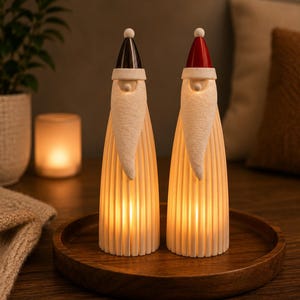 May include: Two tall, conical figurines shaped like Santa Claus with white textured beards and ribbed bodies. One figure features a black hat, while the other has a red hat. Both ornaments are illuminated from within, casting a warm, soft glow.