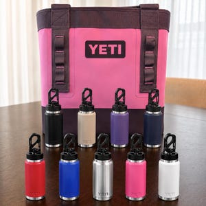May include: A bright pink YETI tote bag with dark trim and a black logo patch, positioned behind nine small cylindrical metal water bottles. Each bottle features a black screw-top lid with a carabiner clip and comes in a different colour.