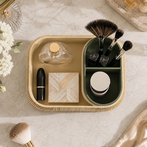 May include: A beige rectangular vanity organiser tray with a ribbed outer edge, featuring a separate dark green compartment for brushes and small items. The tray holds a clear perfume bottle, a black lipstick, a square compact, and brushes.