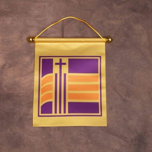 Könnte beinhalten: Ein gelbes Wandbanner aus Stoff mit einem geometrischen Design in Lila und Orange sowie einem zentralen Kreuz. Das Banner hängt an einer goldfarbenen Stange mit dekorativen Endkappen und einer dünnen gelben Kordel zur Aufhängung.