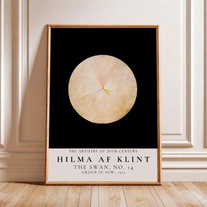 May include: A rectangular art print in a wooden frame featuring a large, pale, circular form against a solid black background. Below the circle, white text reads: THE ARTISTRY OF 20TH CENTURY, HILMA AF KLINT, THE SWAN, NO. 14, GROUP IX SUW, 1915.