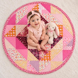 May include: A round patchwork quilt featuring various pink, orange, and yellow floral and geometric fabric patterns. A plush beige rabbit toy with a pink bow sits next to a baby wearing a light pink sleeveless romper and a matching headband.