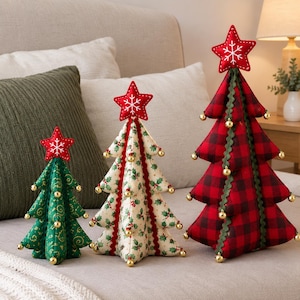 May include: Three fabric Christmas tree decorations in varying sizes. The smallest is green with gold swirls, the middle is cream with holly patterns, and the largest is red and black plaid. Each features a red star topper with a white snowflake.