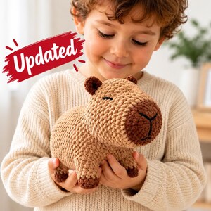 May include: A tan and brown crocheted capybara plush toy with a rounded body, small ears, and a dark brown snout. The toy features a textured yarn surface and is held in place. A red graphic element with the white text Updated is visible.