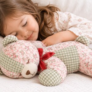 May include: A stuffed teddy bear crafted from fabric featuring a pink floral pattern and a sage green gingham check. The bear has embroidered closed eyes, a brown stitched nose, and a small red ribbon tied around its neck.