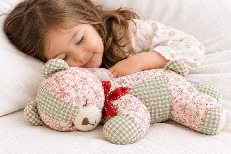May include: A stuffed teddy bear crafted from fabric featuring a pink floral pattern and a sage green gingham check. The bear has embroidered closed eyes, a brown stitched nose, and a small red ribbon tied around its neck.
