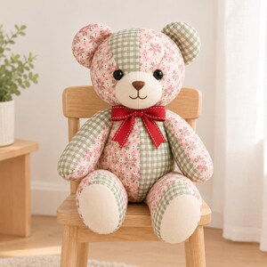 May include: A stuffed teddy bear made of patchwork fabric featuring alternating panels of pink floral print and sage green gingham check. The bear has black button eyes, a brown stitched nose, and a red ribbon bow tied around its neck.
