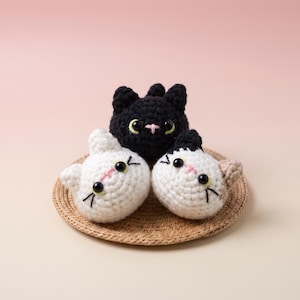 Puede incluir: Tres cabezas de gato pequeñas, redondas y tejidas a ganchillo. Una es negra con ojos amarillos y nariz rosa, mientras que las otras dos son blancas con ojos negros y nariz rosa. Cada cabeza de gato tiene bigotes bordados y orejas pequeñas, dispuestas sobre una alfombrilla redonda tejida.