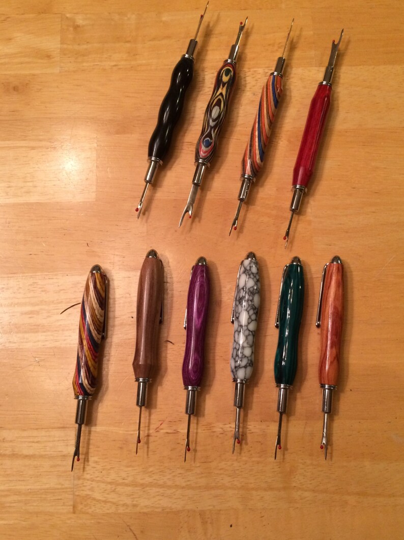 May include: Ten seam rippers with wooden handles in diverse colours and patterns, such as black, red, and striped designs. Each tool features a silver metal blade with a red safety ball. The rippers are displayed on a light brown wooden surface.
