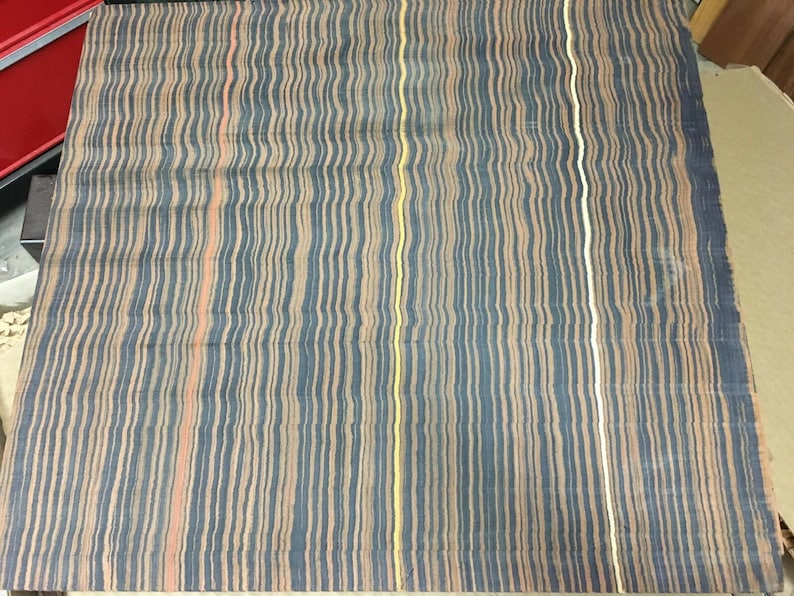 May include: A rectangular textile with a striped pattern. The stripes are vertical and wavy, in shades of blue, tan, and peach, with a few yellow and white stripes. The fabric appears to be folded or slightly wrinkled.