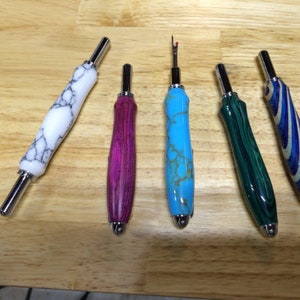 May include: Five seam rippers with decorative handles in various colours. The handles are white with grey marbling, magenta, turquoise with gold veining, green with dark green marbling, and blue and white striped. Each has a silver metal tip.