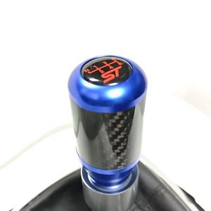 May include: A cylindrical gear shift knob featuring a carbon fiber body with blue metallic top and bottom accents. The top cap displays a black background with a red gear shift pattern and the red text ST. It is attached to a black leather boot.