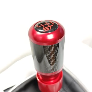 May include: A cylindrical gear shift knob featuring a metallic red top and base with a central black carbon fiber section. The top cap displays a red six-speed manual transmission shift pattern and the letters ST in a bold red font on a black background.