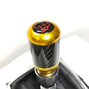 May include: A cylindrical gear shift knob featuring a black carbon fiber body with a metallic gold top and base. The top cap displays a red gear shift pattern and the red text ST. The knob is attached to a black leather shift boot with a silver trim.