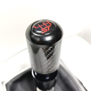 May include: A black cylindrical gear shift knob featuring a carbon fiber pattern on the shaft and a top cap displaying a red manual transmission shift pattern with the letters ST. The knob is attached to a black leather-like shift boot base.