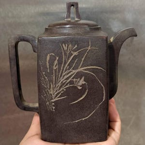 May include: A dark brown, square-shaped ceramic teapot featuring a textured surface and a light-colored, etched floral design on the front. The teapot includes a matching lid with a small handle, a curved spout, and a rectangular side handle.