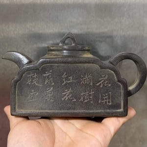 May include: A dark brown, rectangular teapot with a textured surface and a curved handle and spout. The side of the teapot features engraved Chinese characters arranged in two rows, and a small lid with a triangular handle sits on top.