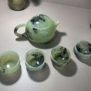 Peut inclure: Un service à thé composé d'une théière ronde avec couvercle et de quatre petites tasses assorties. L'ensemble est fabriqué dans une pierre translucide vert pâle présentant des inclusions sombres, nuageuses et noires marbrées.