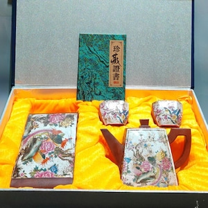 May include: A tea set in a presentation box featuring a teapot, two cups, and a rectangular tray, all decorated with colourful floral and bird patterns. A green booklet with the text 珍藏證書 is included inside the yellow fabric-lined box.
