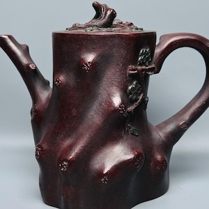 May include: A dark reddish-brown teapot with a textured surface resembling a tree trunk. The teapot features a curved handle, a spout shaped like a branch, and a lid with a small sculpted detail. The body has irregular bumps and knots.