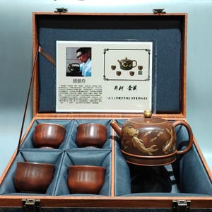 Può includere: Un set da tè in argilla marrone composto da una teiera con decorazioni dorate a forma di carpe koi e quattro tazze abbinate, presentato in una custodia in legno. Il set include una scheda informativa con testo e immagini sulla storia del set.