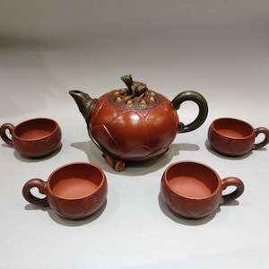 以下が含まれることがあります： 蓋にカエルの彫刻が施され、蓮の花びらのデザインがあしらわれた赤茶色の粘土製ティーポットと、4つの揃いのカップ。ティーポットには濃い緑色の取っ手と注ぎ口があり、底には小さな蓮の種子の鞘のアクセントが付いています。