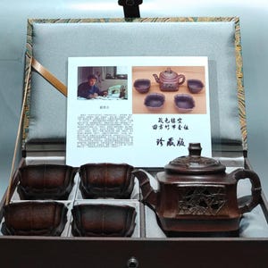 Könnte beinhalten: Ein dunkelbraunes Ton-Teeservice mit einer quadratischen Teekanne mit bambusartigen Griffen und vier passenden Tassen, präsentiert in einer stoffgefütterten Box. Eine Informationskarte mit Bildern und chinesischem Text liegt im Deckel.