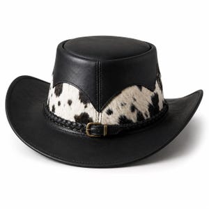 May include: A black leather hat featuring a wide brim and a flat crown. The base of the crown is accented with a band of white and black cowhide fur. A thin, braided black leather strap with a small brass-toned metal buckle encircles the hat base.