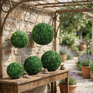 May include: A collection of six artificial boxwood topiary balls in varying sizes. Two spheres are suspended from the overhead metal structure by ropes, while four spheres are arranged on the surface of a wooden table. The foliage is deep green.
