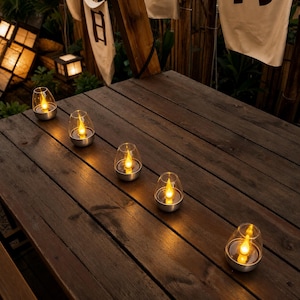 May include: Five small, clear glass lanterns with silver-toned bases are arranged on a dark brown wooden table. Each lantern contains a warm, glowing LED light shaped like a flickering flame, casting a soft illumination across the wooden planks.