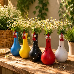 May include: Five ceramic vases shaped like chickens, each featuring a wide base and a narrow neck. The vases are colored blue, yellow, black with white dots, red with black dots, and white with black dots. Each holds a bouquet of white daisies.