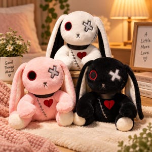 以下が含まれることがあります： 白、ピンク、黒の3体のウサギのぬいぐるみ。各ウサギには、黒い目、十字型の目、胸に縫い付けられたハート、黒い首輪が付いています。ウサギは長い耳をしており、体には縫い目のディテールが施されています。