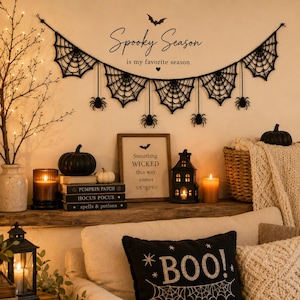 May include: A black crocheted spider web garland with hanging spider accents, displayed on a wall above a shelf. The wall features the text Spooky Season is my favorite season and a framed sign reading Something WICKED this way comes.