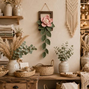 May include: A wall-mounted rectangular dark wood frame featuring a large, crocheted pink flower with a cream center and a long, trailing vine of dark green crocheted leaves hanging downward from the bottom of the frame.