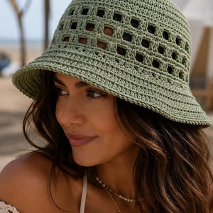 May include: A sage green crochet bucket hat featuring a dense, solid crown and a wide, open-work square grid pattern around the mid-section. The hat has a short, slightly downturned brim with a reinforced edge, crafted from textured yarn.