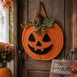 May include: A round, orange crocheted jack-o'-lantern wall hanging with a brown stem, green leaves, and curly vines. The pumpkin features a black crocheted face with triangular eyes, a triangular nose, and a wide, toothy, grinning mouth.