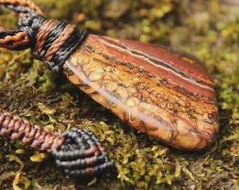 May include: A pendant featuring a polished, multi-colored stone with swirling patterns of brown, tan, and cream, suspended from a braided cord made of black and brown fibers. The stone has a smooth, glossy surface and an irregular, elongated shape.