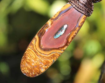 May include: An elongated, polished pendant featuring a central reddish-brown stone with swirling golden-brown patterns and a small, iridescent, blue-green inlay at the center, suspended from a dark brown, braided cord necklace.