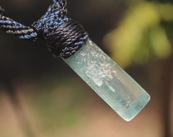 May include: A cylindrical, pale blue, translucent crystal pendant suspended from a braided cord. The cord is crafted from black and blue fibers, tightly wrapped around the top of the stone to secure it in place, creating a textured, woven appearance.